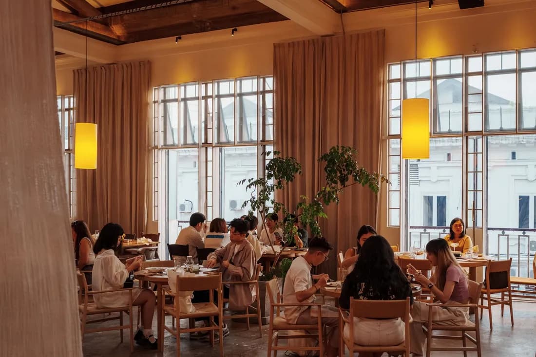 Bright, airy restaurant dining room with guests at wooden tables in warm evening light